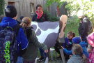 Eine Gruppe Kinder steht vor einer Holzkuh, die Erlebnisbäuerin steht dahinter, am rechten Bildrand versuchen sich zwei Kinder am Melken