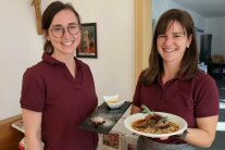 Zwei Frauen in rotem Polo-Shirt präsentieren Teller mit Speisen