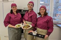 Drei Studierende zeigen Vorspeise, Hauptspeise und Nachspeise in der Schulküche.