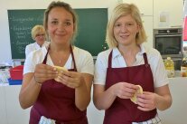 Vier Frauen mit weißem Oberteil und roter Schürze stehen hinter verschiedenen Backformen.