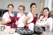 Vier Frauen mit weißem Oberteil und roter Schürze stehen hinter verschiedenen Backformen.