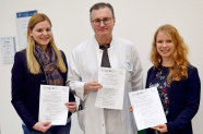 In der Mitte steht ein Mann mit weißem Kittel und links und rechts von ihm zwei Frauen. Alle halten ein weißes Papier in der Hand