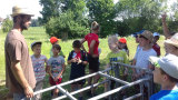 Kinder stehen gemeinsam mit dem Bauern und der Lehrerin um eine Abferkelbucht